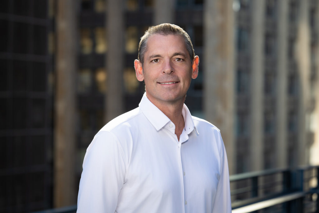 Professional on-location headshot of male executive photographed outdoors with natural light and blurred urban architecture background for corporate branding and LinkedIn profile. For Headshot Photography Portfolio