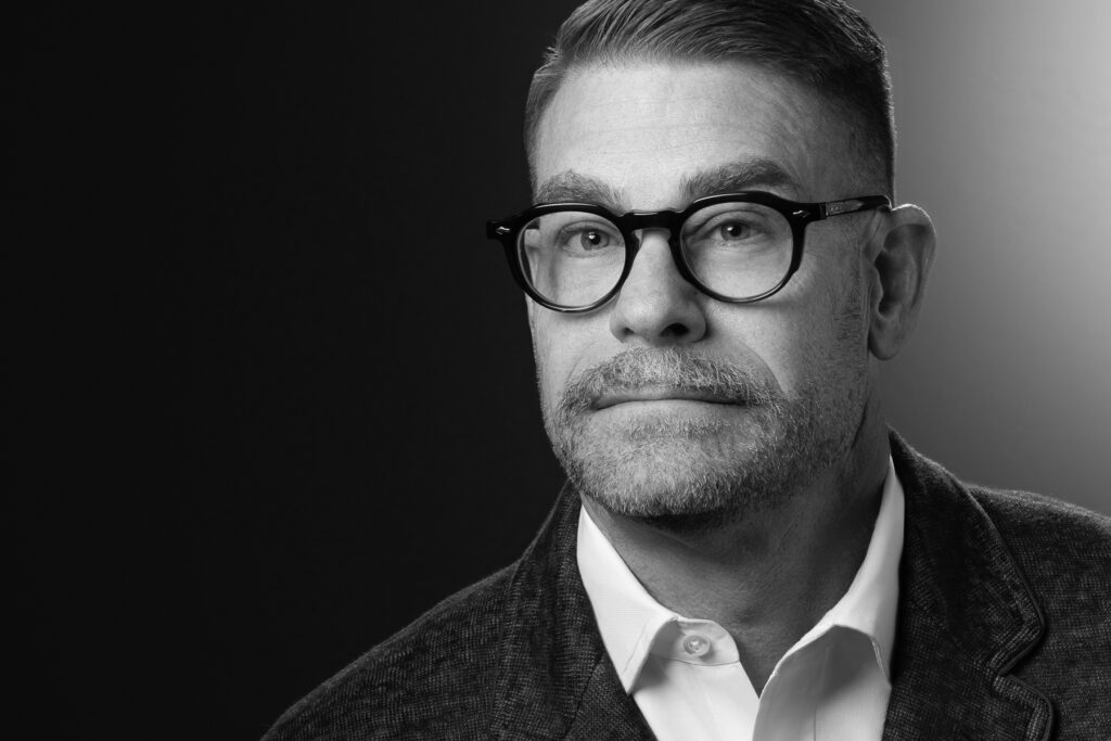 Black and white studio headshot of male executive wearing glasses photographed with dramatic studio lighting and dark gradient background for corporate branding and professional profile