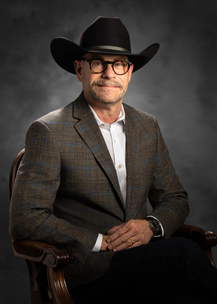 Professional studio portrait of male executive wearing cowboy hat and glasses seated in chair photographed with dramatic studio lighting and textured dark background for executive branding and corporate profile