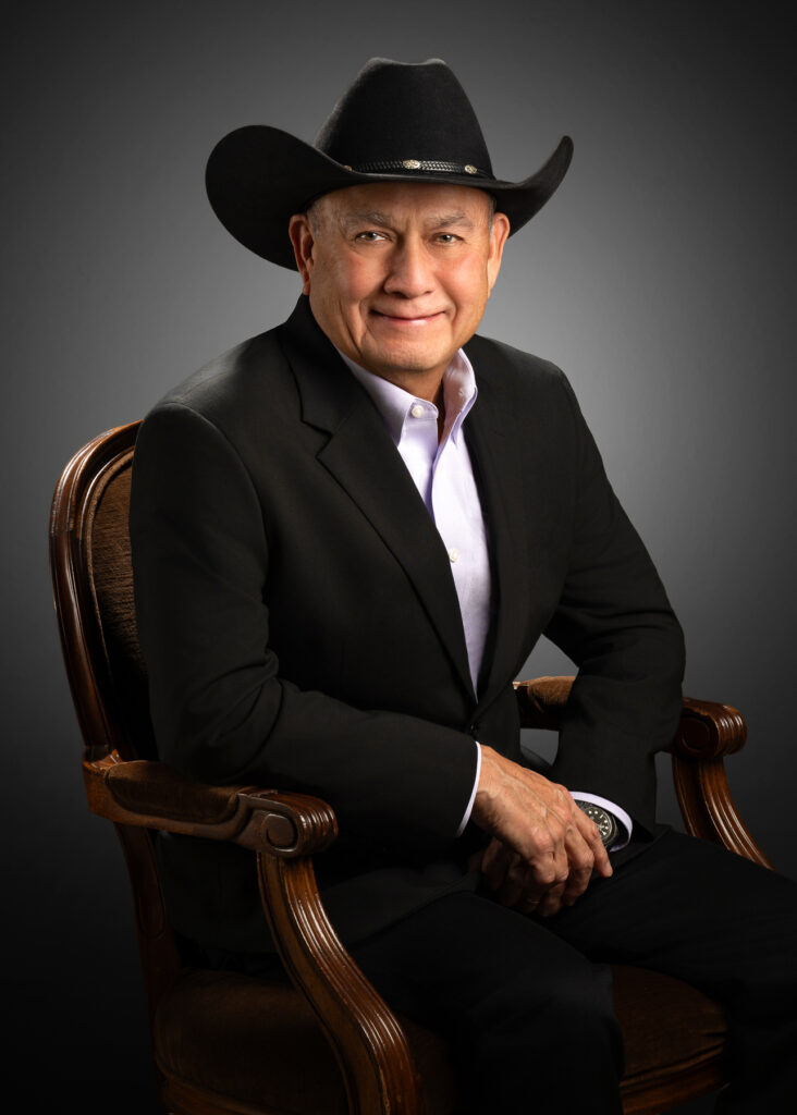 Professional studio portrait of senior male executive wearing black cowboy hat and suit seated in chair photographed with dramatic studio lighting and dark gradient background for corporate branding and executive profile