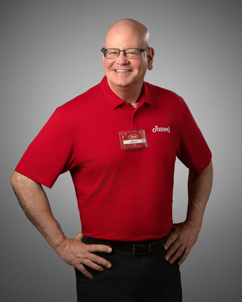 Professional studio headshot of retail employee wearing red branded company polo shirt and name badge photographed against neutral gray studio background for corporate staff directory and branding