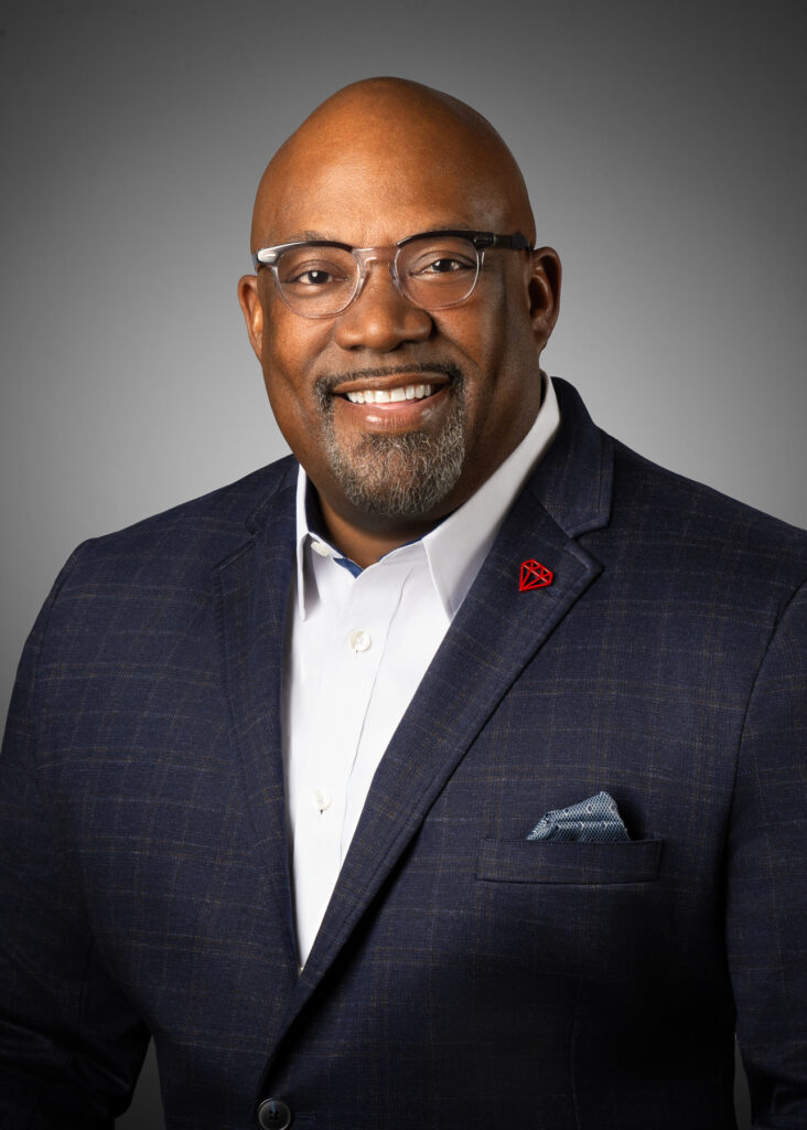 Professional studio headshot of male executive wearing glasses and navy blazer photographed with soft studio lighting and neutral gray gradient background for corporate branding and LinkedIn profile