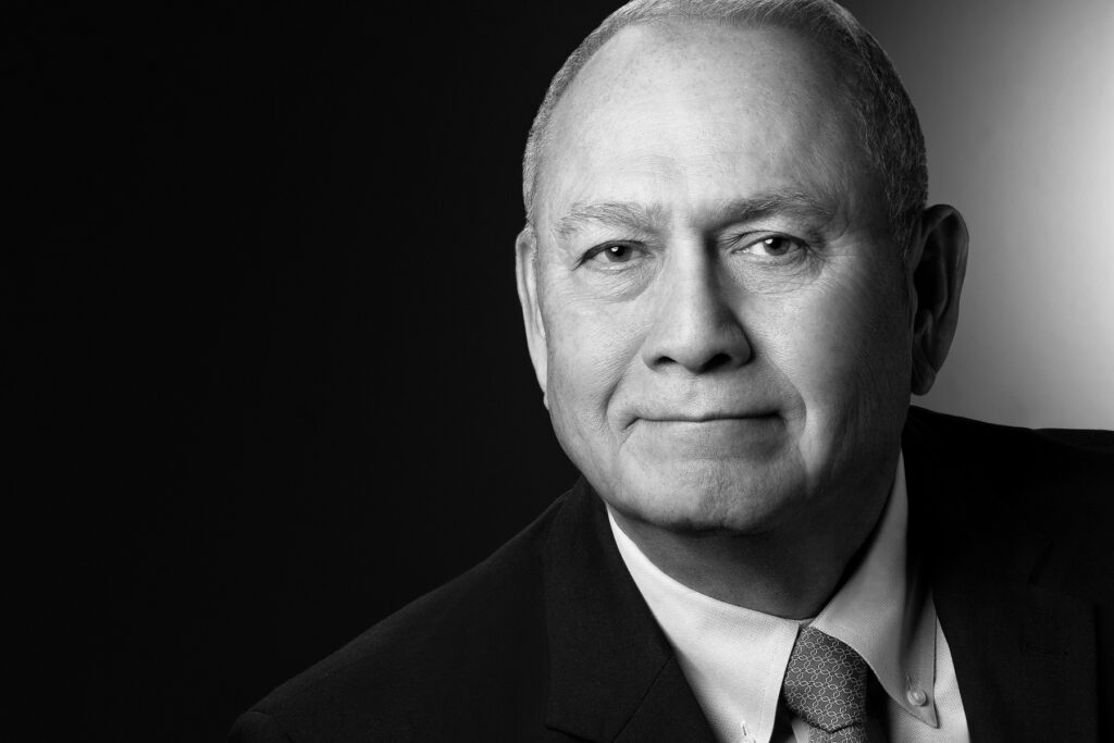 Black and white studio headshot of senior male executive wearing suit and tie photographed with dramatic studio lighting and dark gradient background for corporate branding and professional profile
