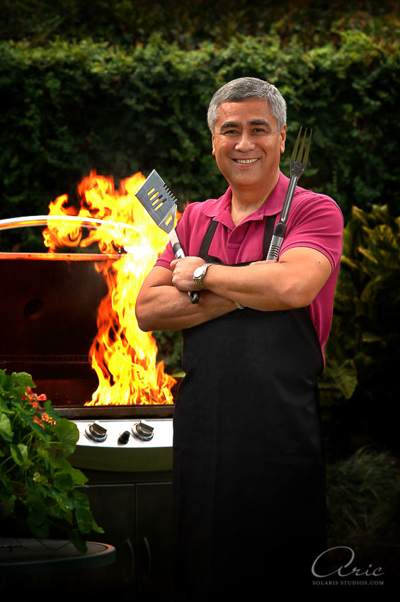 Portrait of Bill Balleza standing beside a flaming grill, holding barbecue tools and smiling for a summer editorial cover image.