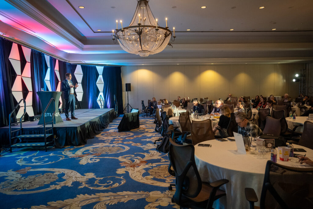 Corporate event breakout session with a speaker presenting on stage to attendees seated at round tables in a ballroom.