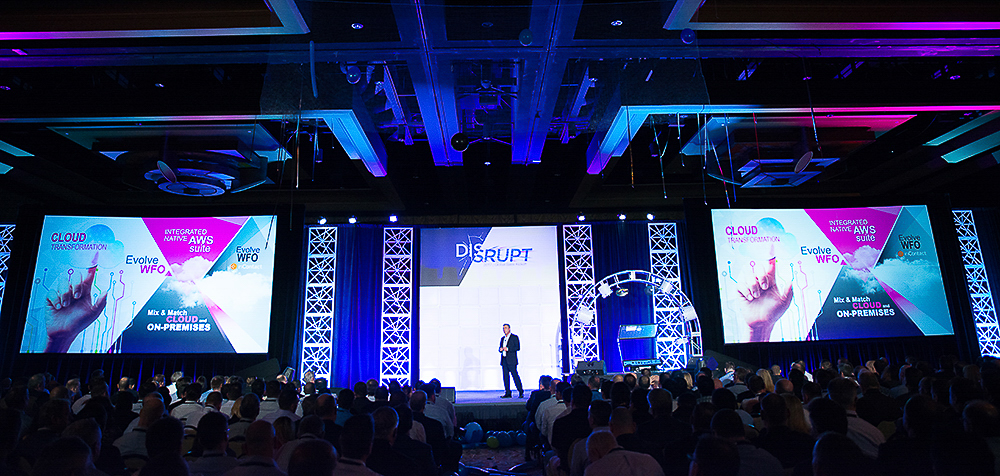 Wide rear-of-room view of a large corporate event audience facing a speaker on stage at Hilton Americas in Houston.