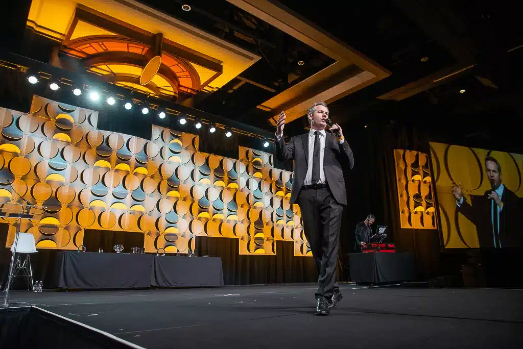 Low-angle stage photo of a keynote speaker presenting at a large corporate event in Houston with dramatic lighting and a patterned stage backdrop.