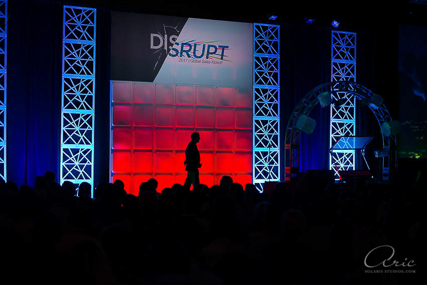 Silhouette of a speaker walking across a brightly lit stage during a corporate event, with the audience visible in the foreground.