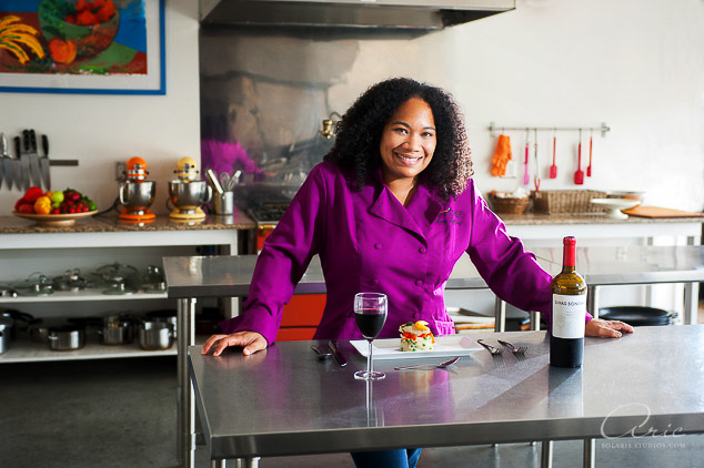 Portrait of a chef in a magenta jacket standing in a commercial kitchen beside a plated dish and a glass of red wine.