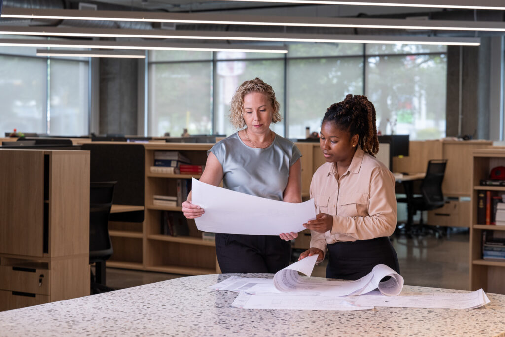 Editorial office portrait of two employees reviewing plans inside an engineering firm in Austin, with a wide view of the workspace and balanced window light.