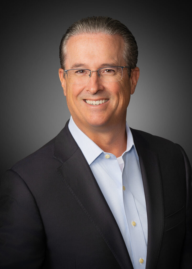 In-studio business headshot of a man in a dark jacket and open-collar shirt against a gray vignette background.