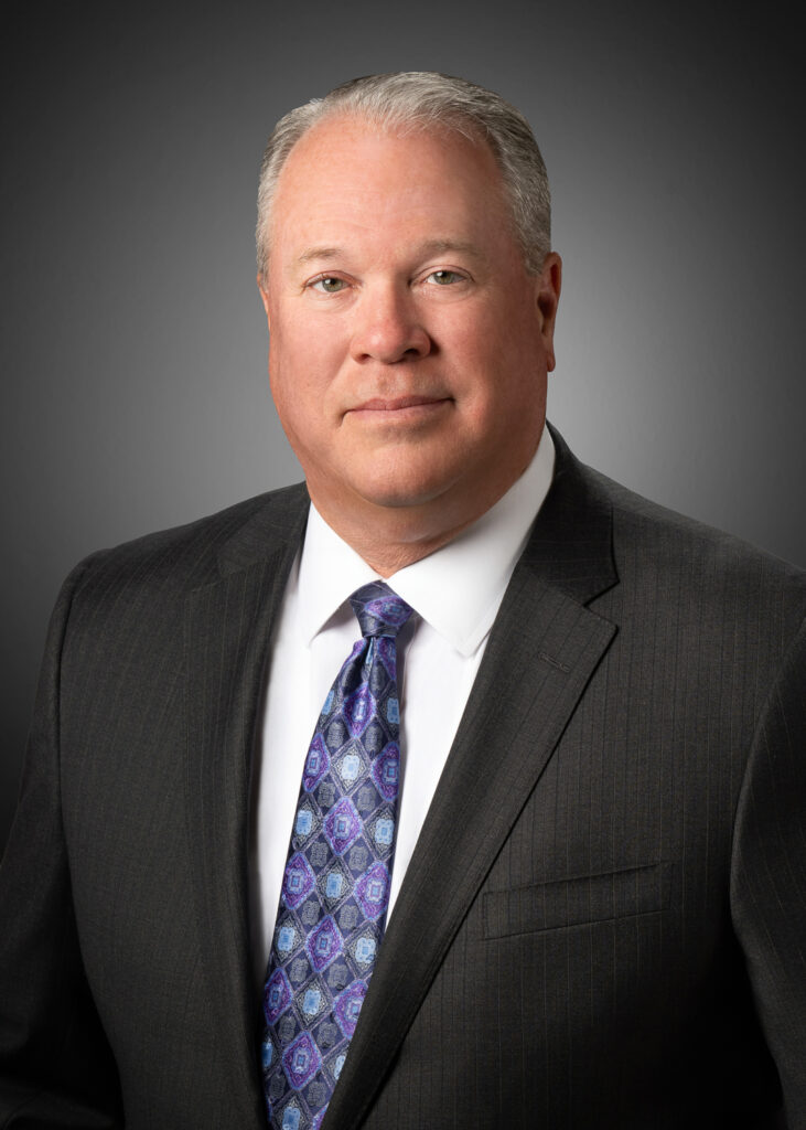 In-studio corporate headshot of a businessman in a dark suit and patterned tie against a gray vignette background.
