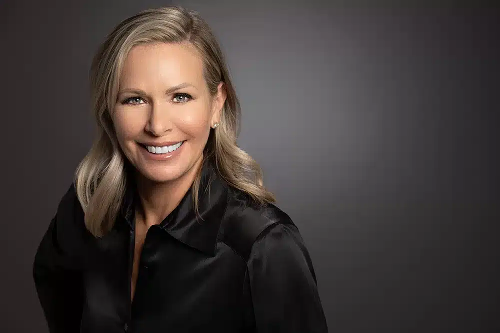 Professional business headshot of a smiling woman in a black blouse against a dark gray studio background