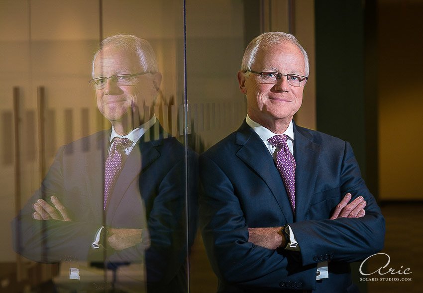Executive portrait of a businessman in a dark suit standing beside a glass wall, with his reflection visible in an office interior.