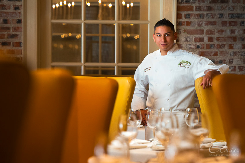 Portrait of the executive chef at Brennan’s Houston standing in the dining room, framed by yellow chairs and warm interior light.