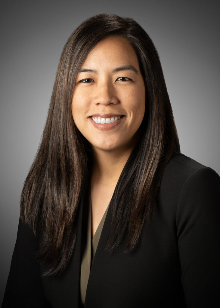 Classic in-studio headshot of a professional woman in a dark blazer against a gray vignette background.