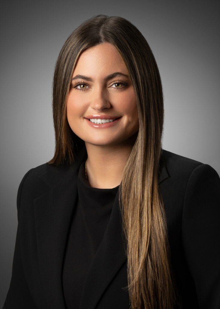 Classic in-studio headshot of a professional woman in a black blazer against a gray vignette background.