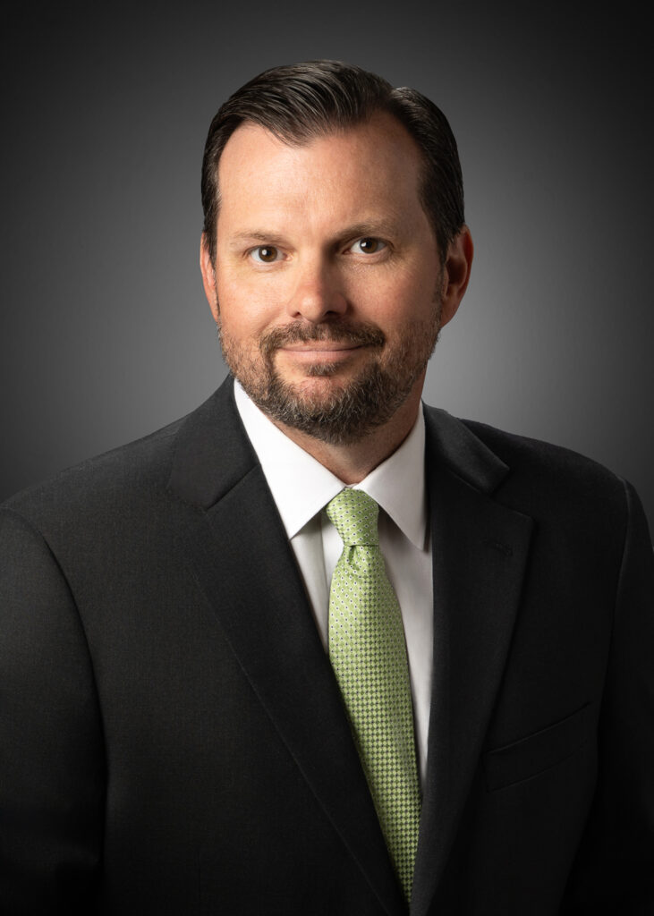 Classic in-studio headshot of a businessman in a dark suit and light green tie against a gray vignette background.
