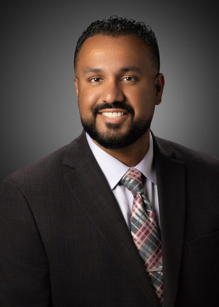 Professional in-studio headshot of a businessman in a dark suit and patterned tie against a gray vignette background.