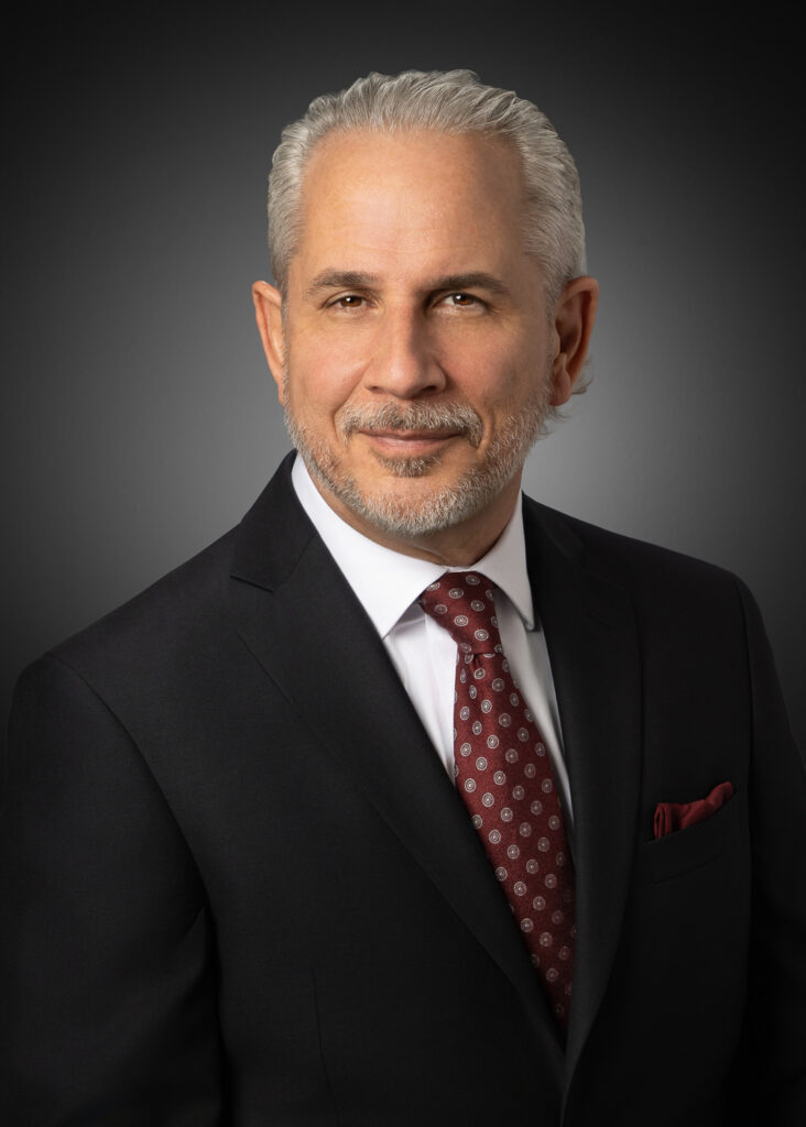 In-studio executive headshot of a businessman in a dark suit and red tie against a gray vignette background.