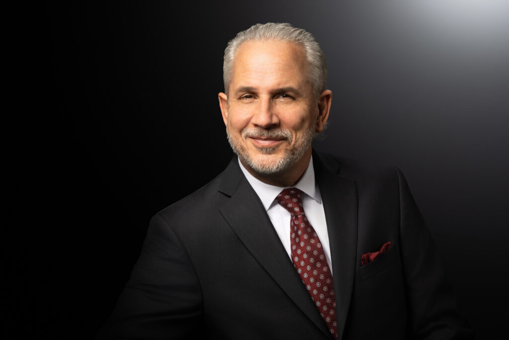 Horizontal in-studio headshot of a businessman in a dark suit and red tie, positioned slightly off-center against a black-to-gray gradient background.