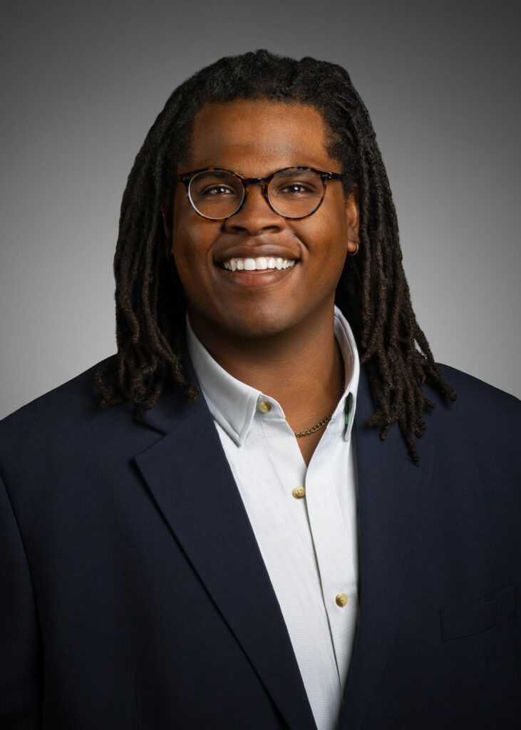 In-studio headshot of a smiling businessman with glasses and shoulder-length hair, photographed against a gray vignette background.