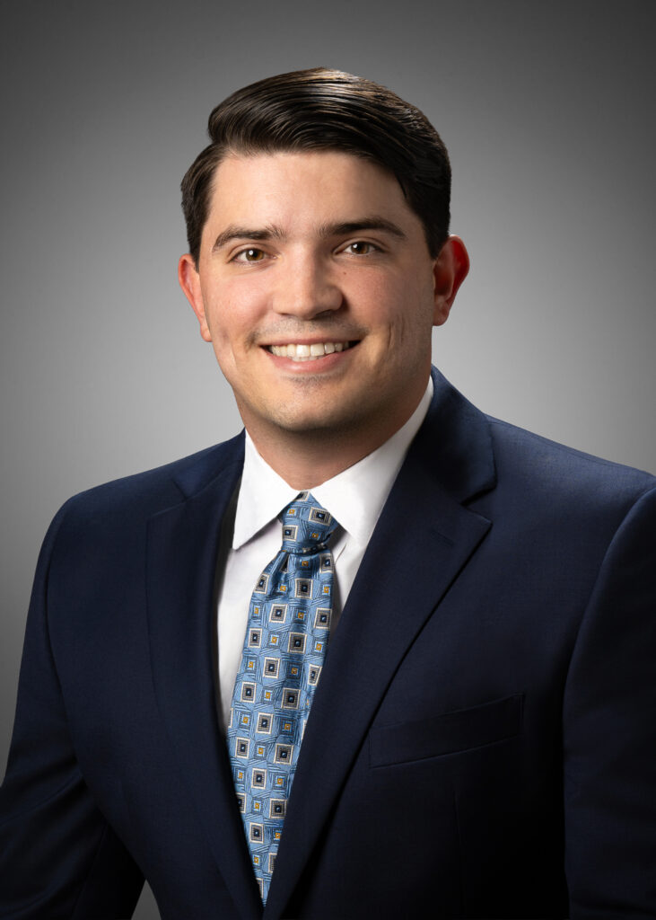 In-studio headshot of a young businessman in a blue suit and patterned tie against a gray vignette background.