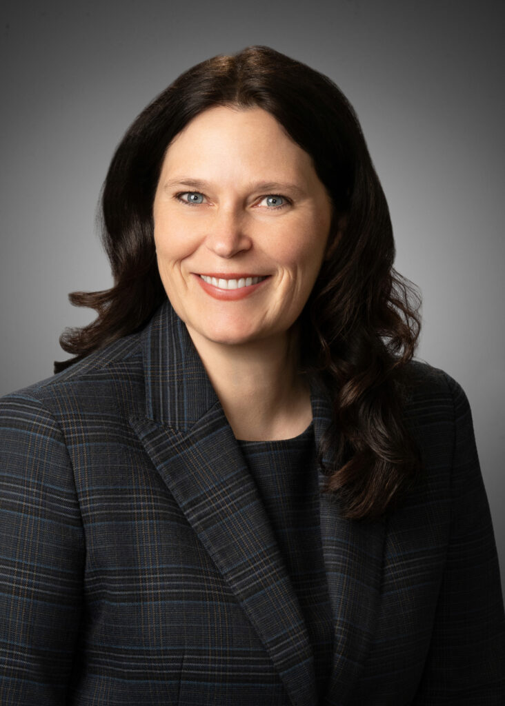In-studio headshot of a professional woman in a dark plaid jacket against a gray vignette background.