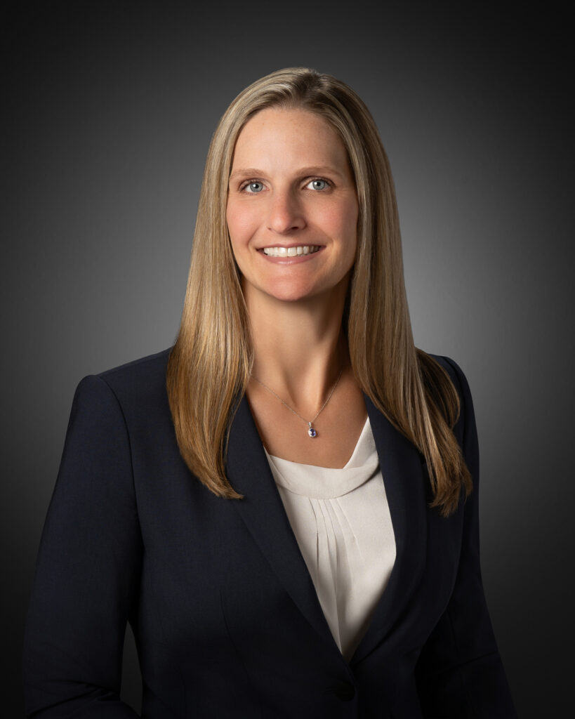 In-studio headshot of a professional woman in a navy blazer and white blouse against a gray vignette background.