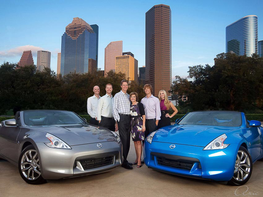 Family portrait of the Baker Nissan team posed between two Nissan sports cars with the downtown Houston skyline behind them at sunset